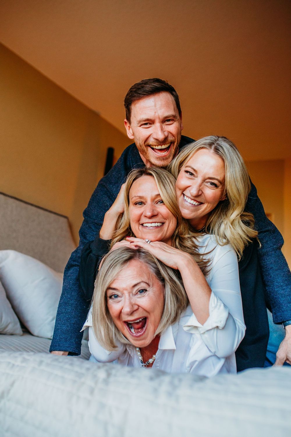 Four smiling adults playfully stacked on a bed, laughing together in a cozy bedroom setting.