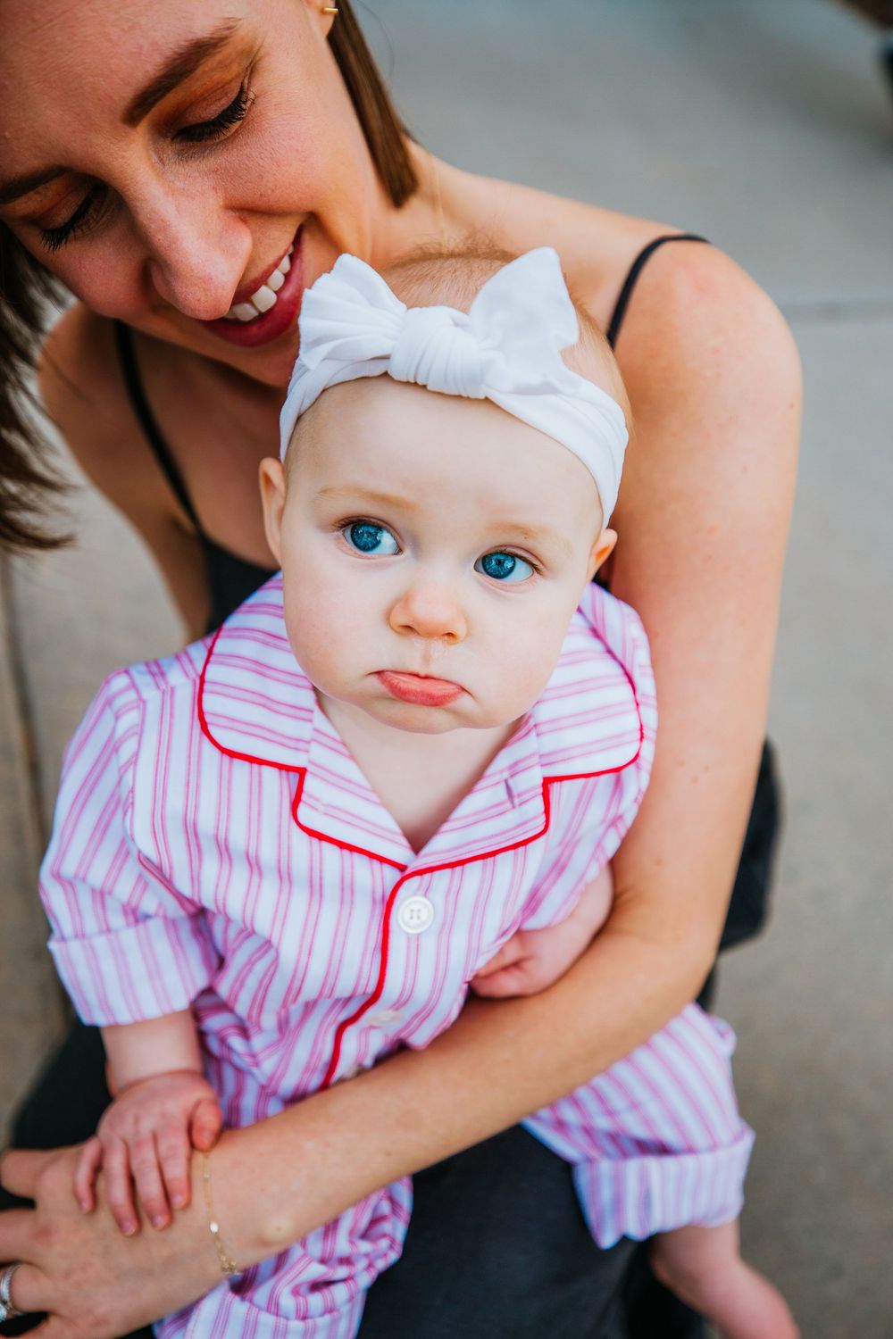 A smiling mother holds her blue-eyed baby girl wearing a pink striped outfit and white bow headband.