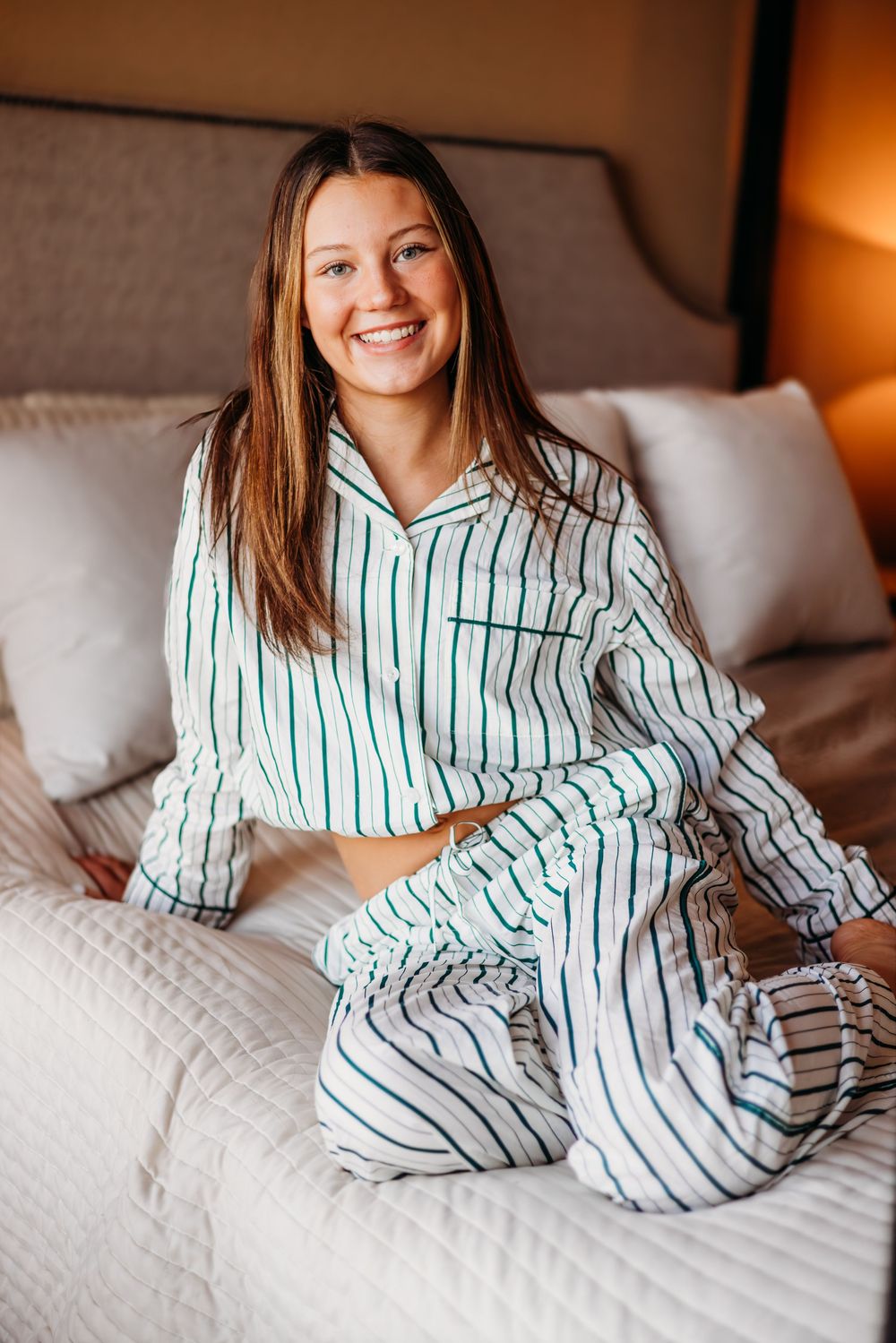 Smiling young woman sitting on bed wearing white and green striped pajamas in cozy bedroom.