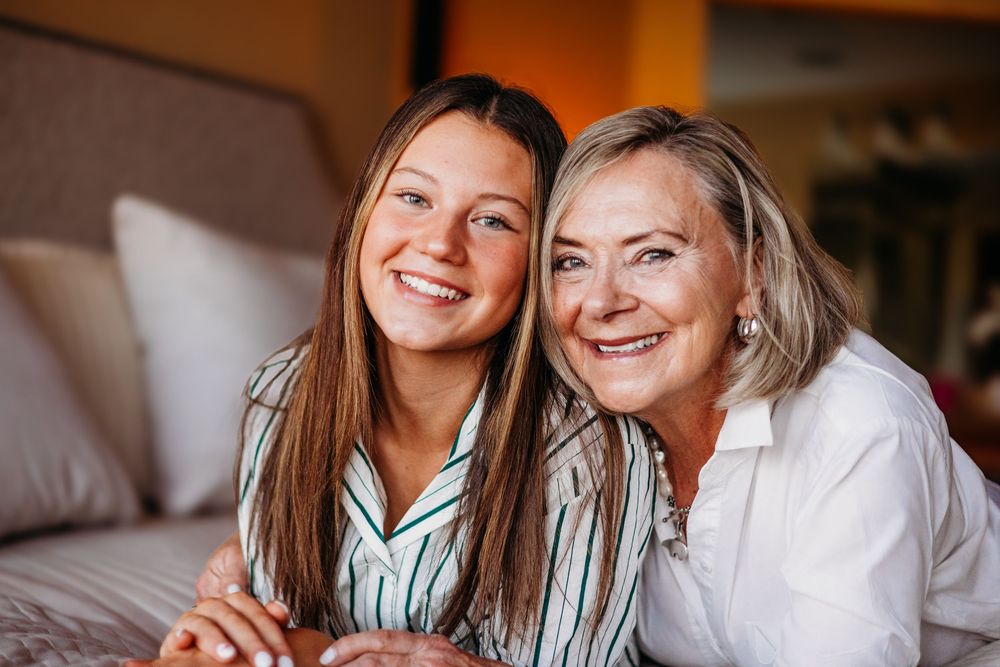 Two smiling women, likely mother and daughter, pose together on a bed in a warmly lit bedroom.