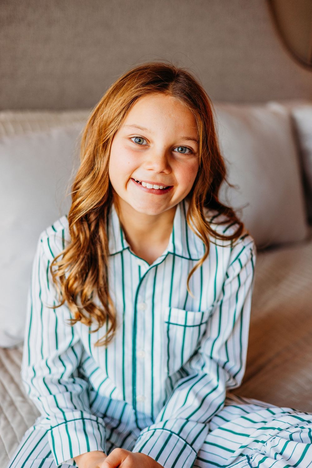 Smiling young girl with red hair wearing white and green striped pajamas sitting on a bed.