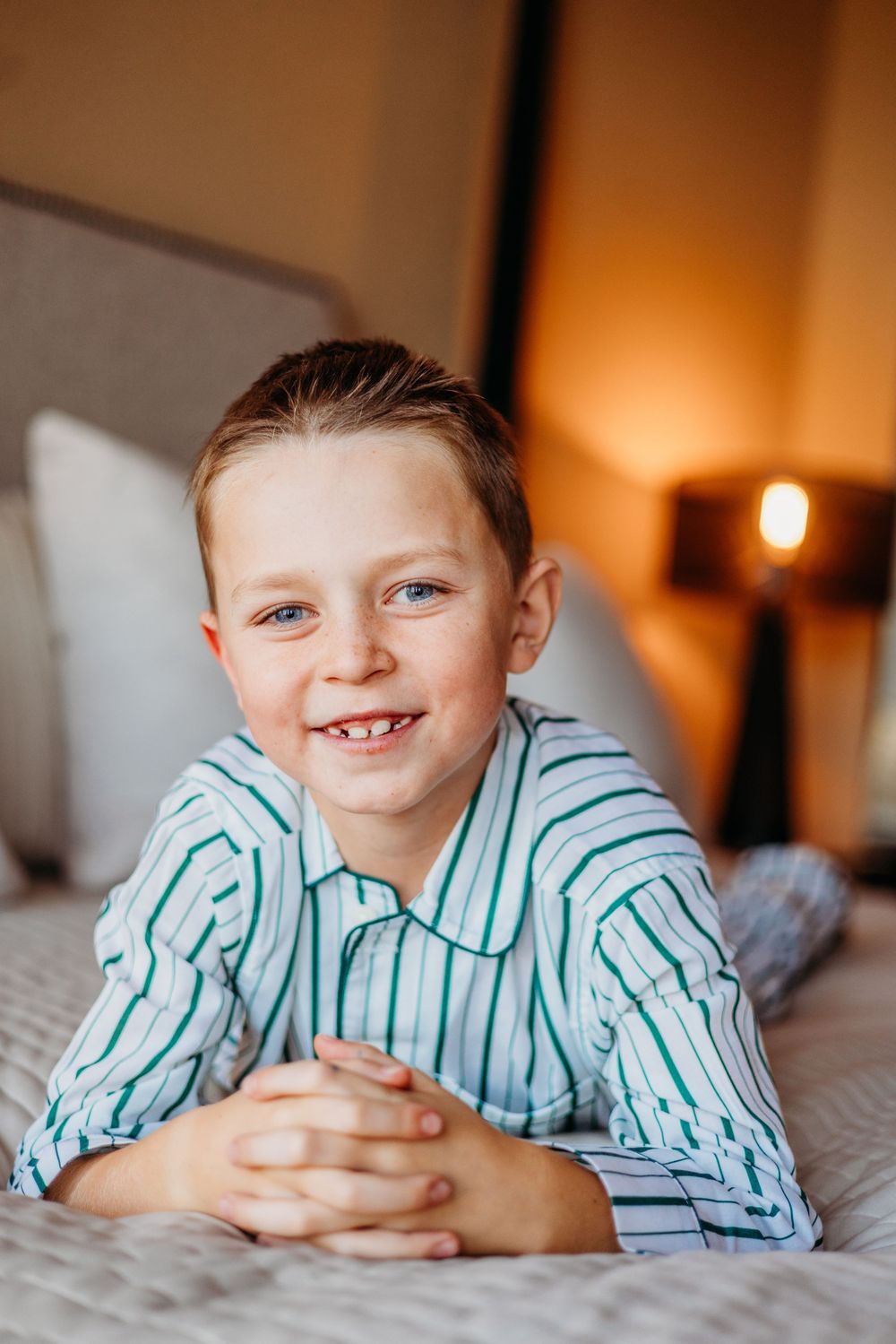 Smiling boy in striped blue pajamas lying on bed with hands clasped, warm lamp glowing in background.