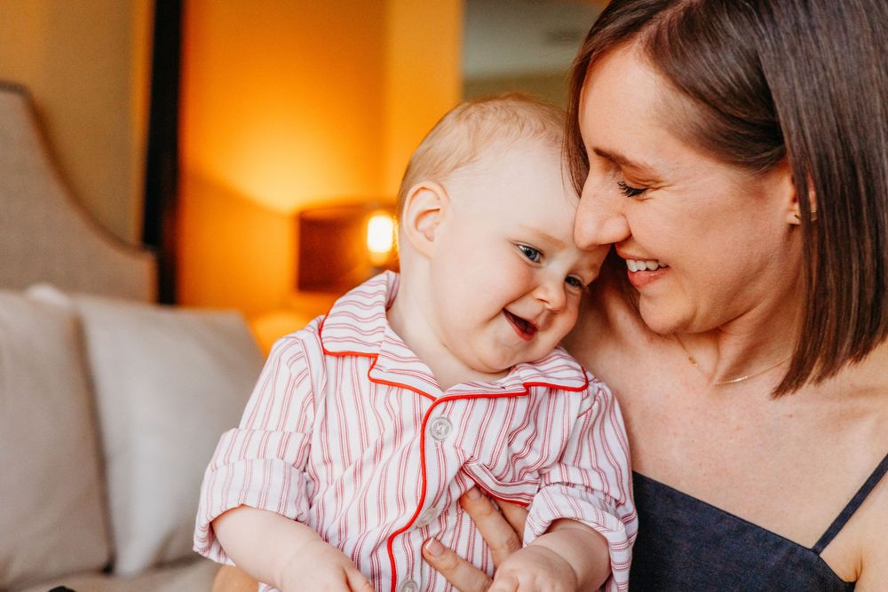A joyful baby in striped pajamas laughs while being held by a smiling brunette woman in a warmly lit room.