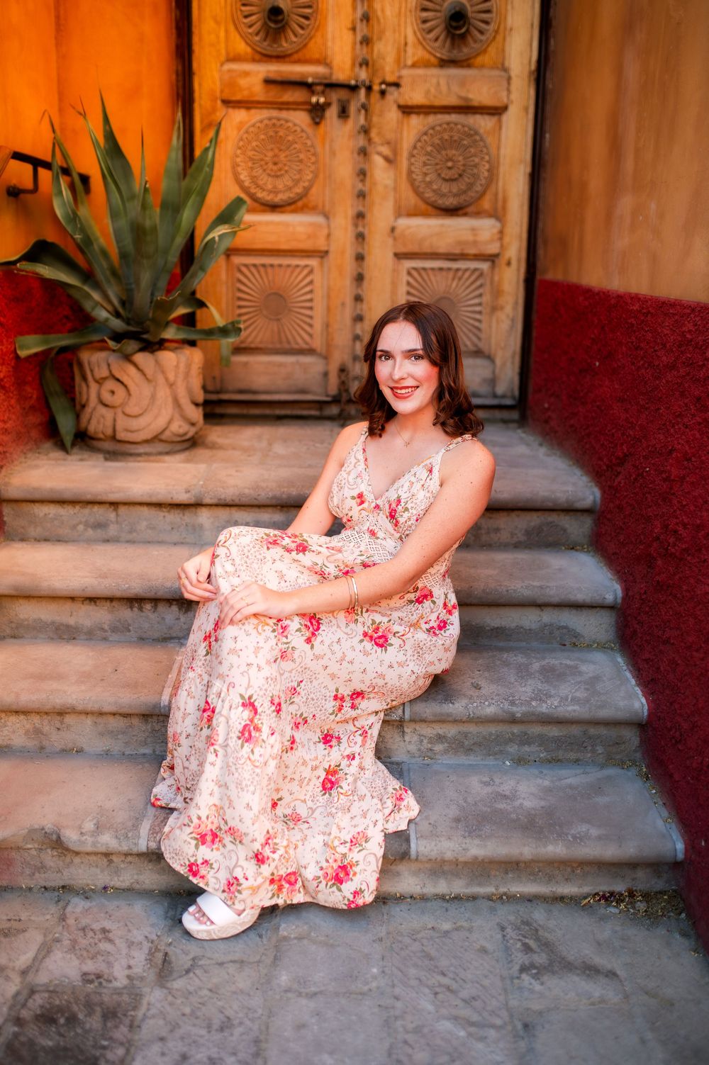Woman in floral maxi dress sitting on stone steps by ornate wooden door with potted plant.