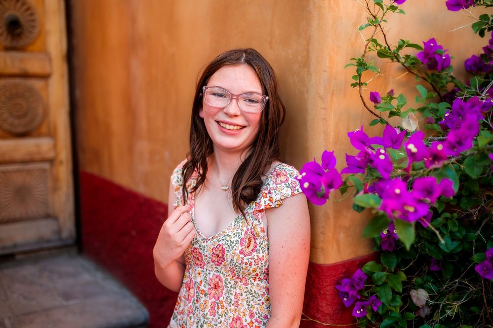 Young woman in floral dress and glasses smiling beside purple bougainvillea flowers against a colorful wall.