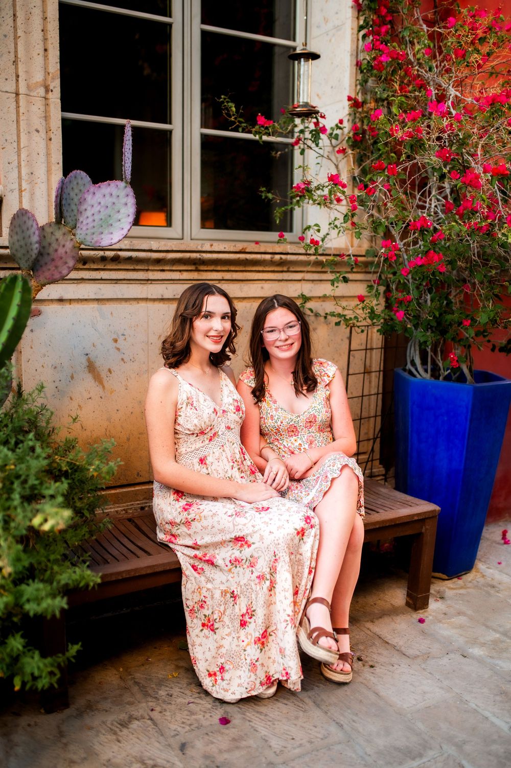 Two young women in matching floral dresses sitting on a wooden bench surrounded by colorful plants and flowers.