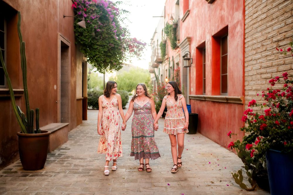 Three women in floral dresses walk hand-in-hand through a colorful alley with red walls and blooming flowers.