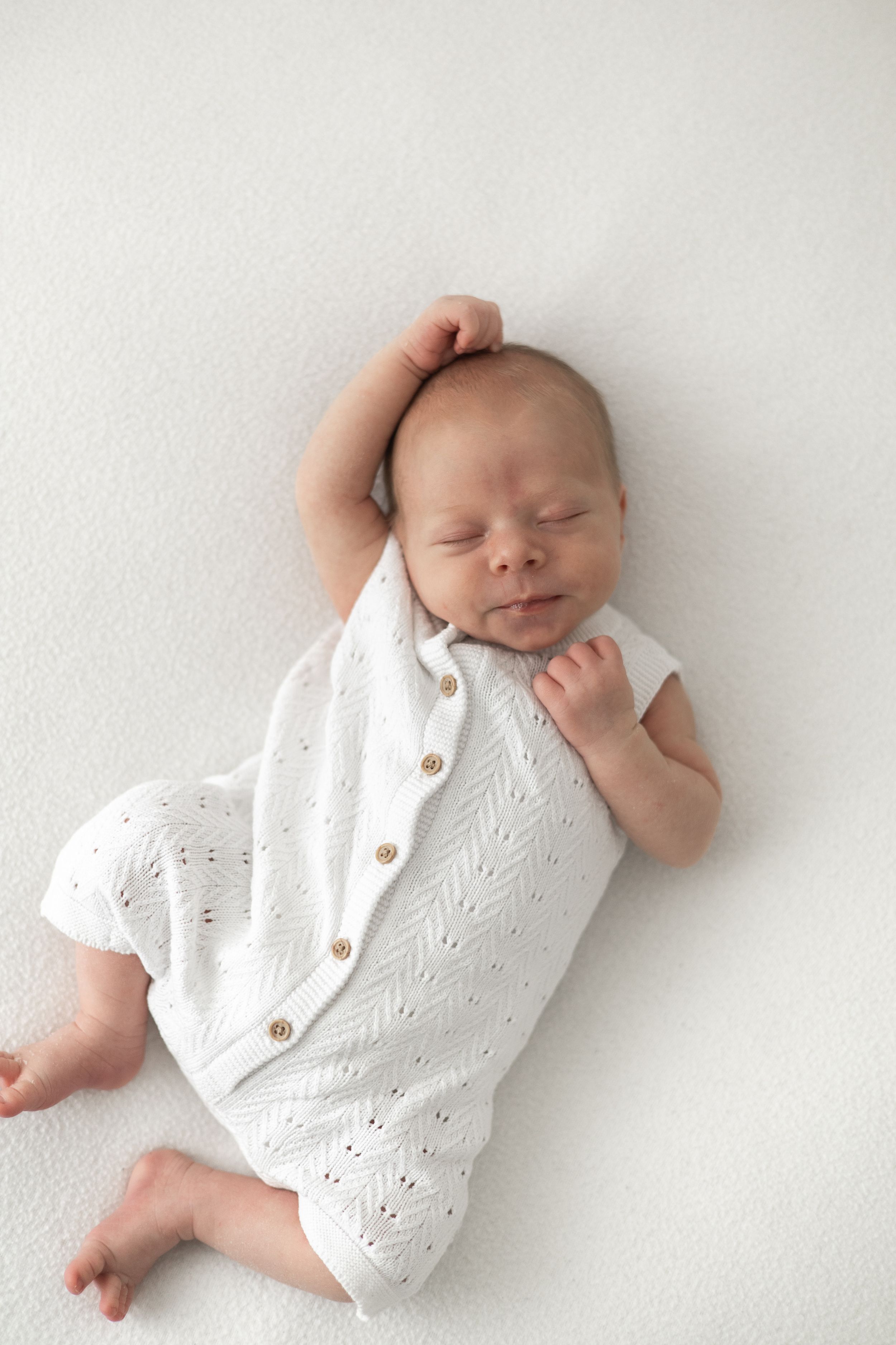 Peaceful newborn baby sleeping in white knit romper on white background, arms raised above head Nottingham newborn photographer