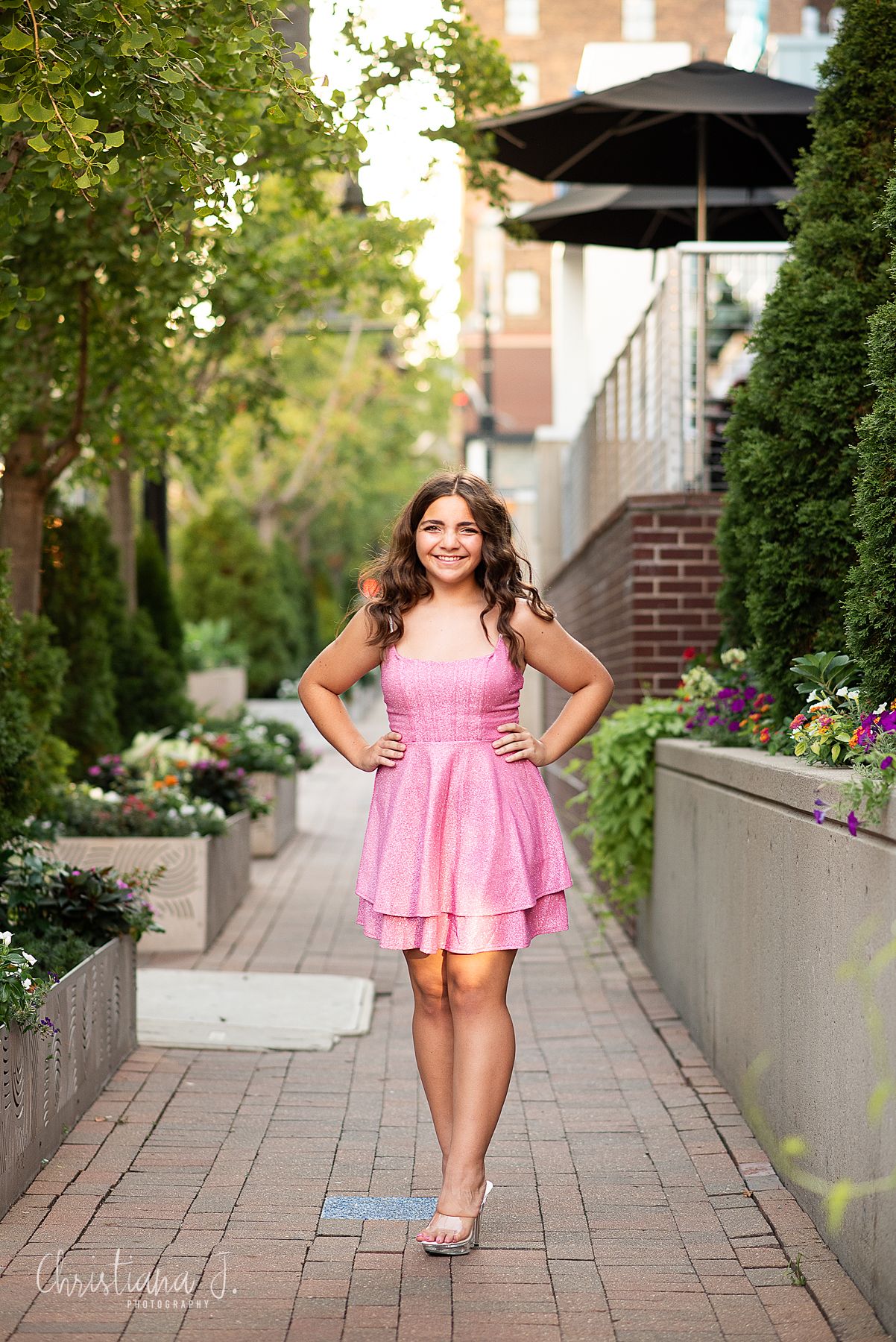 Young woman in pink tiered dress posing on a brick pathway surrounded by greenery.