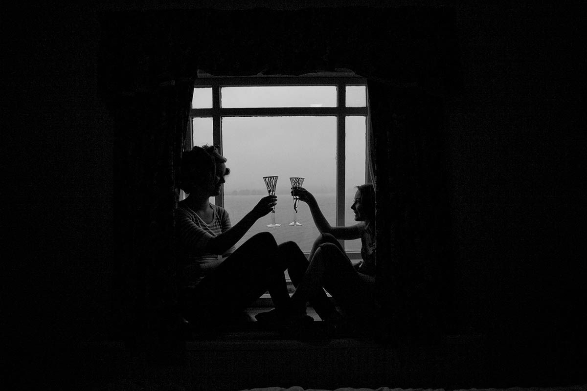 Two people silhouetted against a window toasting with wine glasses in a moody black and white photo.