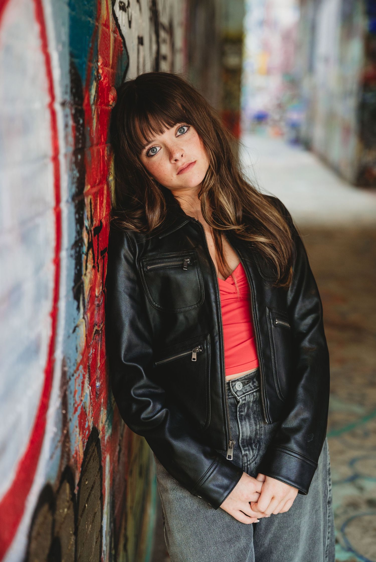 Young woman in black leather jacket and red top leaning against graffiti wall in urban alley.