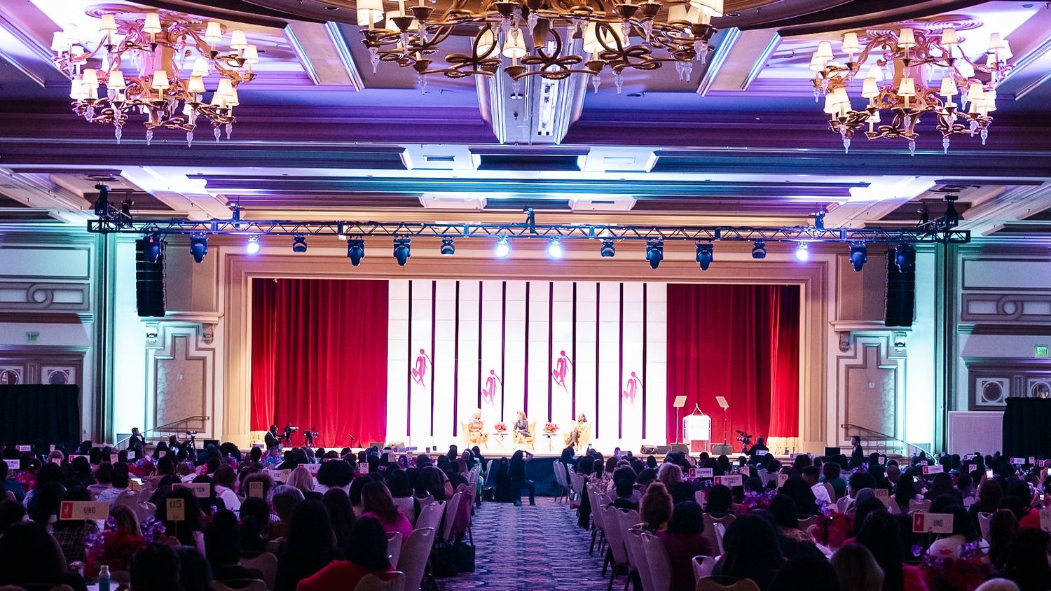 Elegant ballroom event with chandelier ceiling, packed audience facing a red-curtained stage with colorful lighting.
