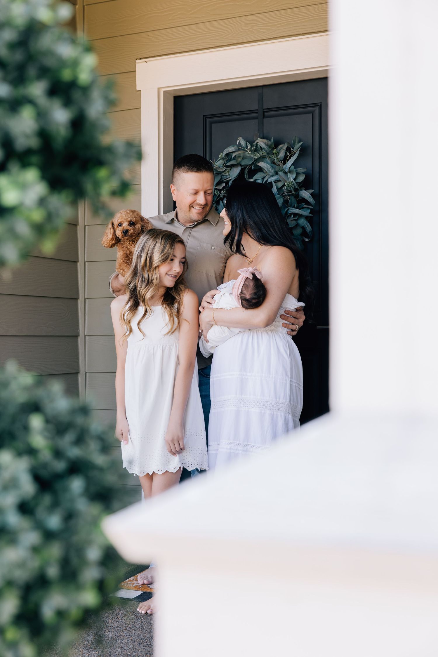 Family of four with a dog poses at their front door, mom holding newborn, daughter in white dress.