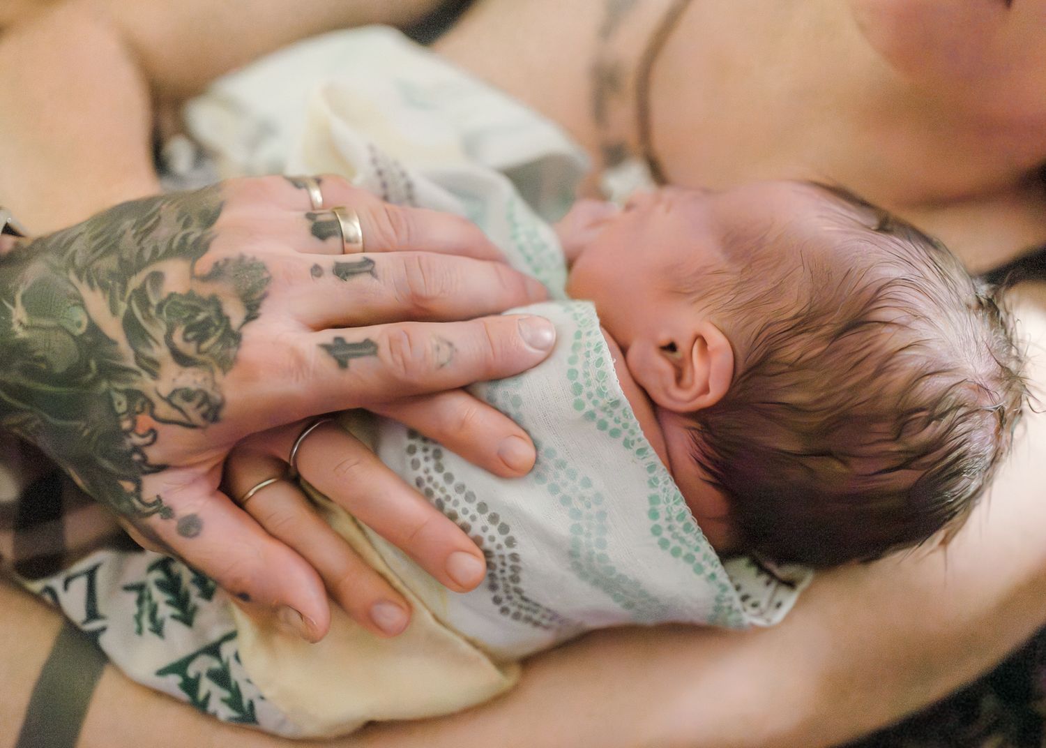 Tattooed hands cradling a sleeping newborn baby wrapped in a soft blanket against a mother's chest.