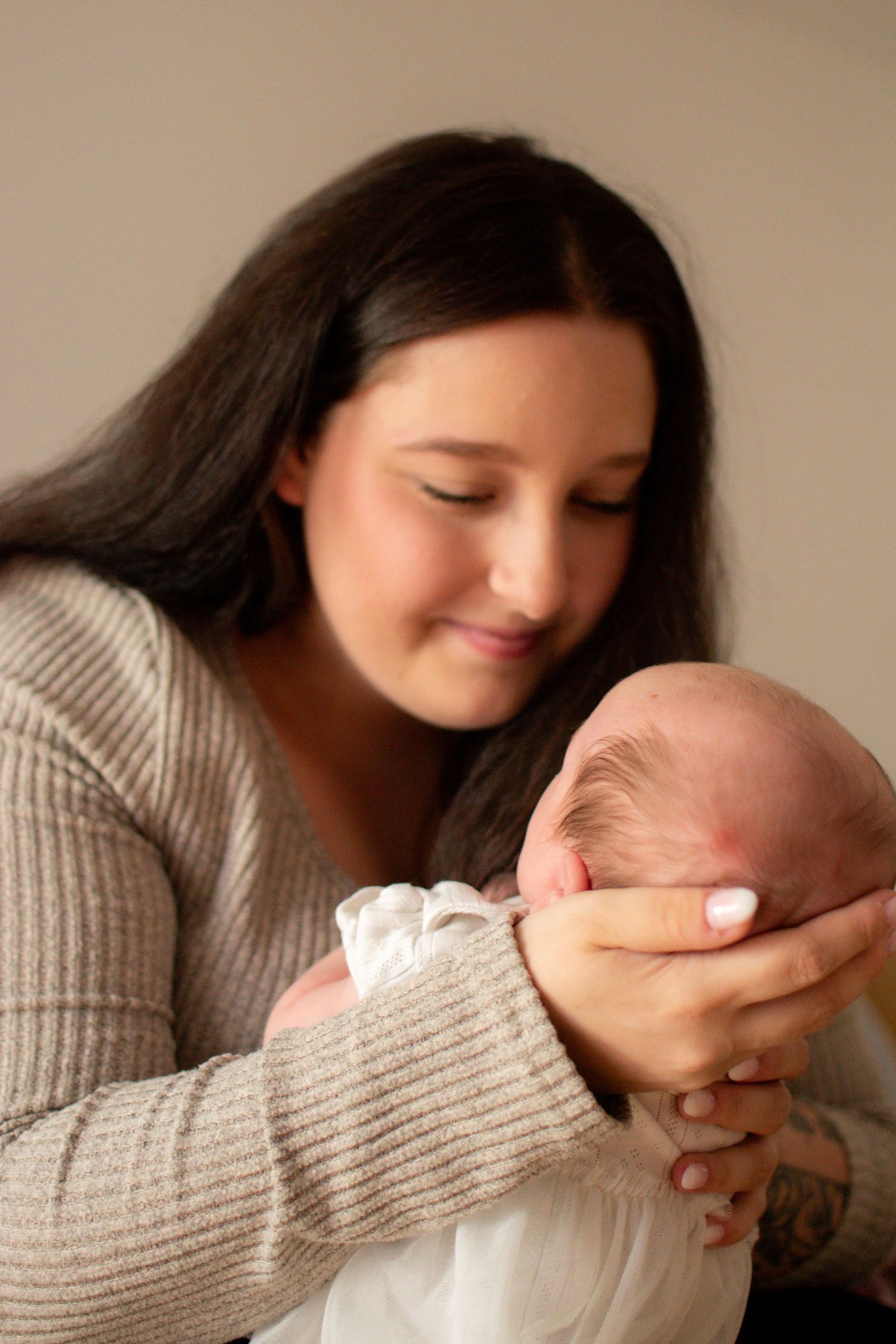 A young dark-haired mother in a beige sweater tenderly holds her newborn baby, gazing down with a gentle smile.