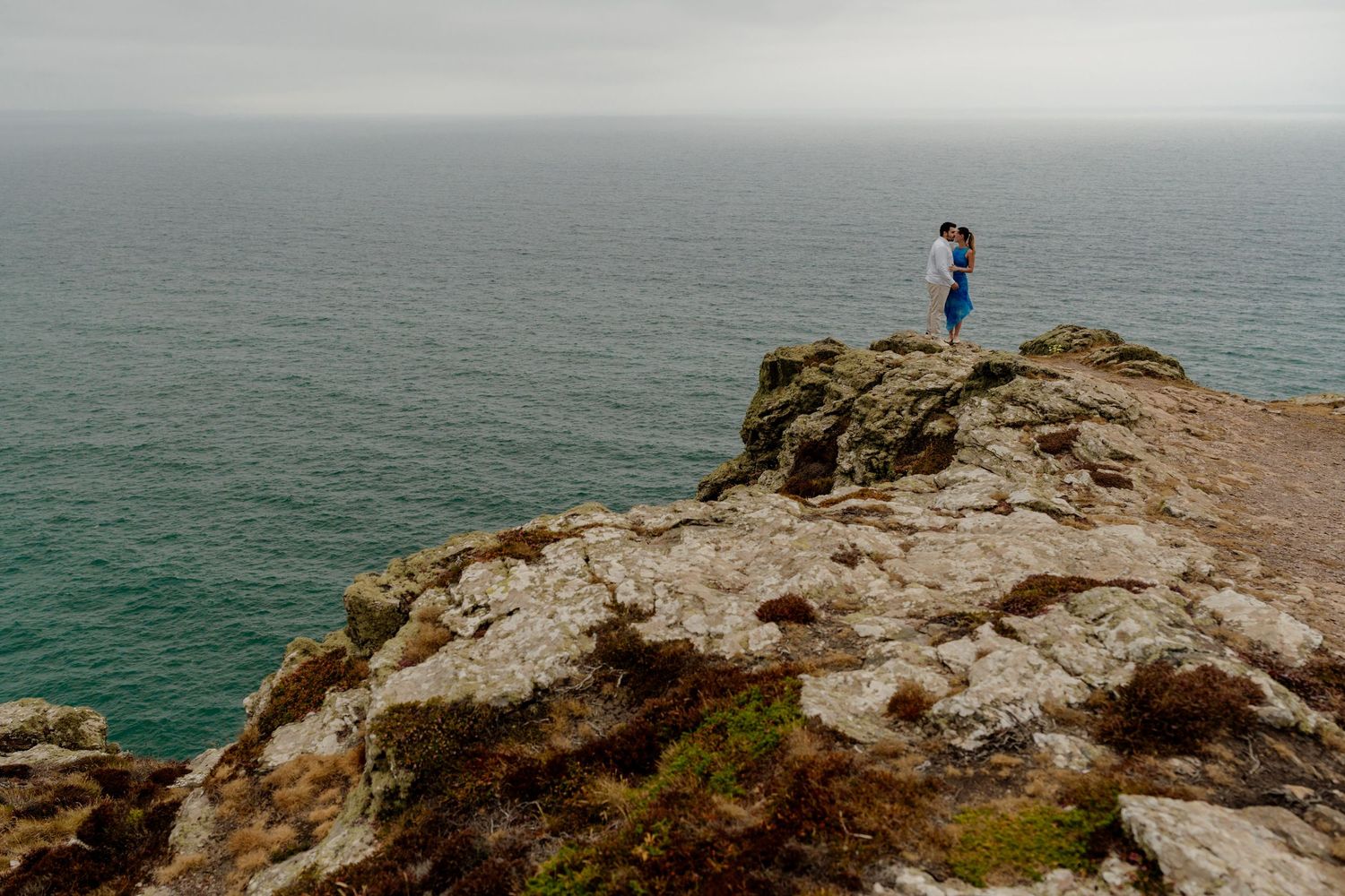 Couple embracing on st agnes head cliffs overlooking a vast turquoise ocean under an overcast sky.