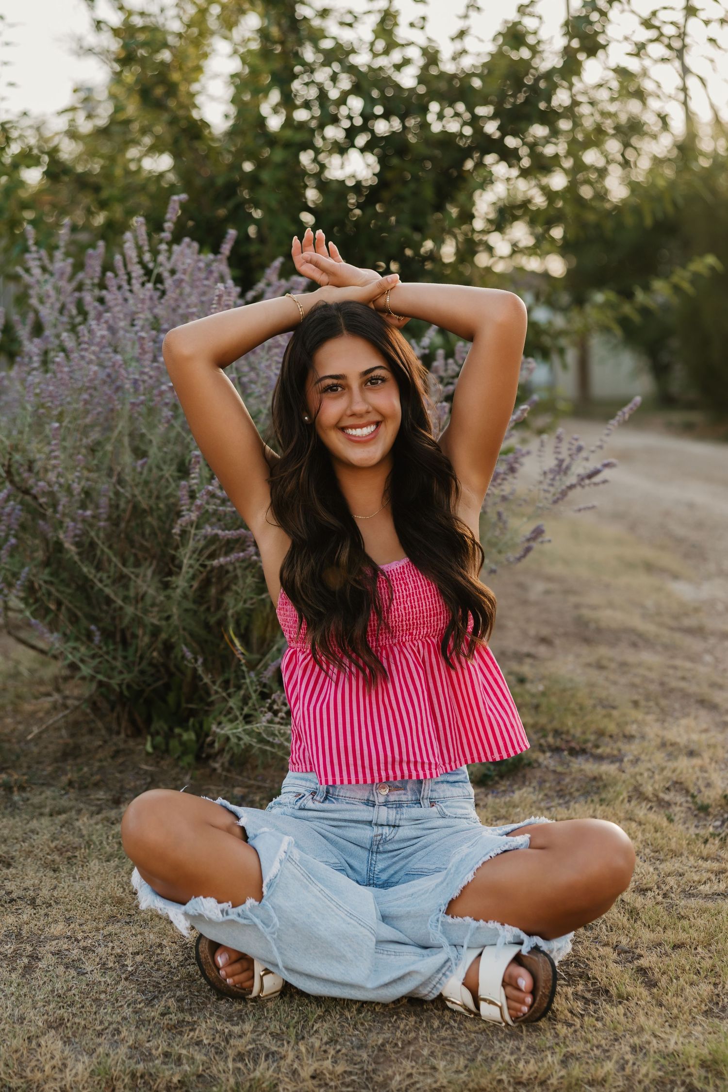 A senior girl smiles with arms over head for a dramatic pose wearing a fun pink top and ripped jeans in front of sage bush in Tulia Texas
