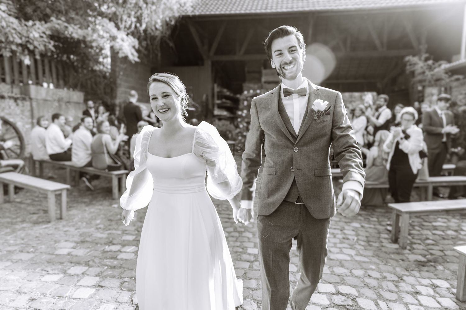 Happy newlywed couple walking hand in hand in black and white wedding photo at outdoor venue.