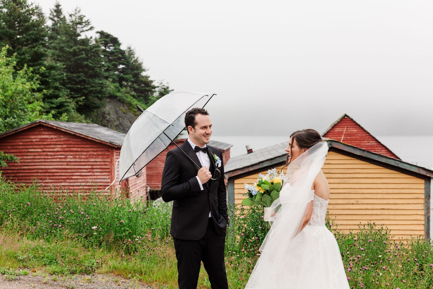 Bride and groom share a romantic moment outdoors holding a clear umbrella near rustic red and yellow buildings by the water.