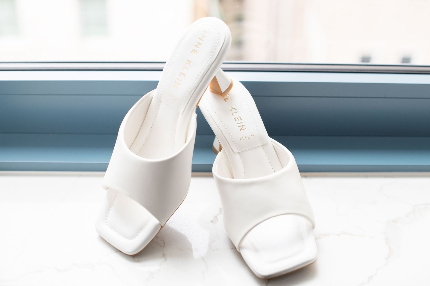 White square-toe mule heels displayed on a marble surface near a window, perfect bridal or wedding shoes.
