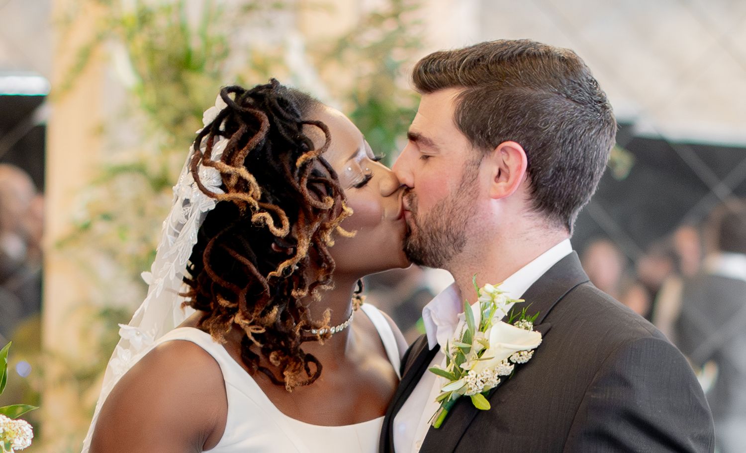 Interracial couple sharing a first kiss at their wedding ceremony, bride in white dress, groom in gray suit.