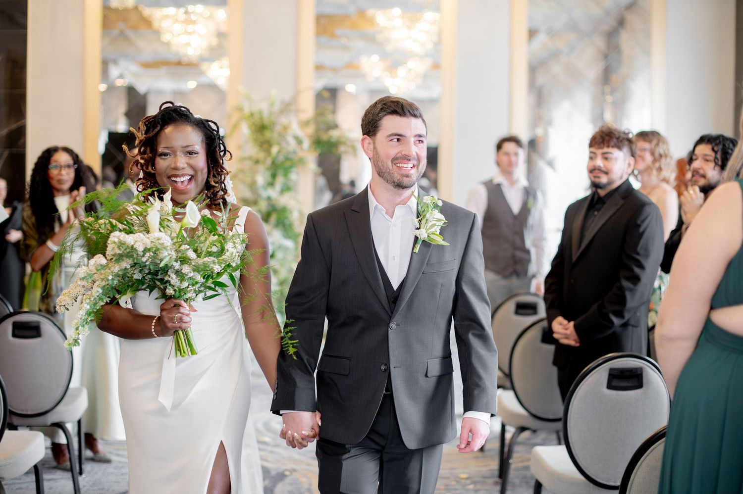 Joyful interracial couple walks down wedding aisle, bride holding white bouquet, groom in gray suit, guests applauding.