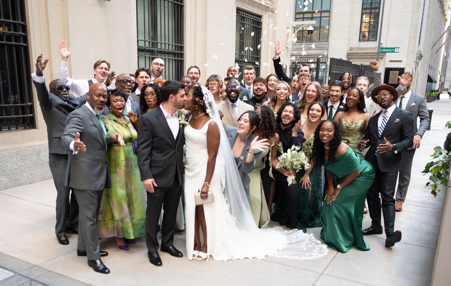 Wedding party celebrating outdoors, bride in white gown with bouquet, guests in green dresses and suits, cheering joyfully.