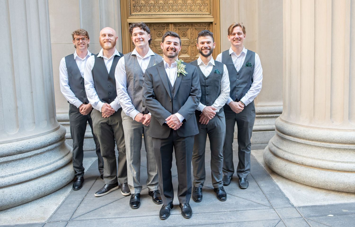 Groom and groomsmen in gray suits and vests posing on steps of elegant building with stone columns.