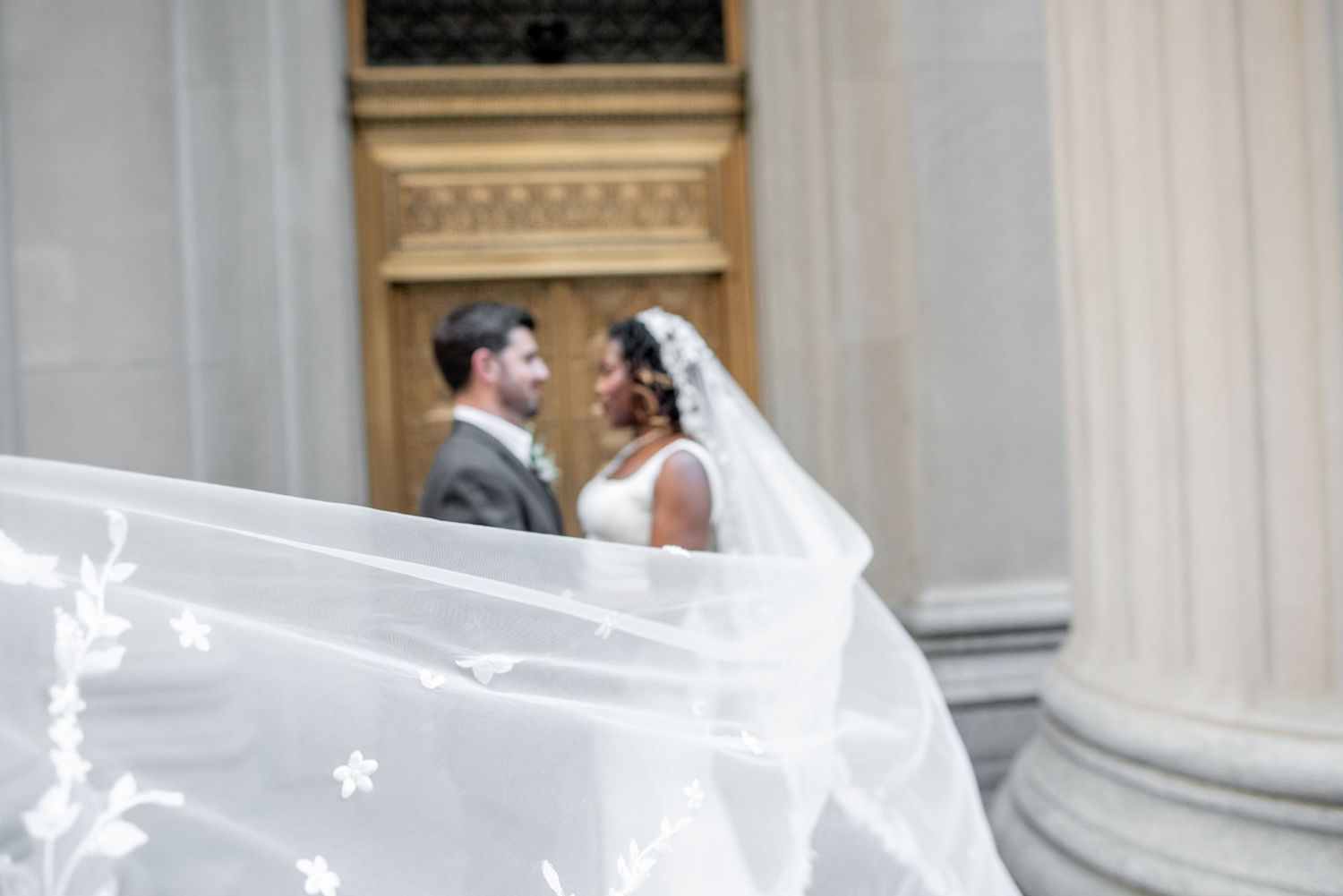 Bride and groom share a romantic moment on grand stone steps, her floral veil billowing gracefully in the wind.