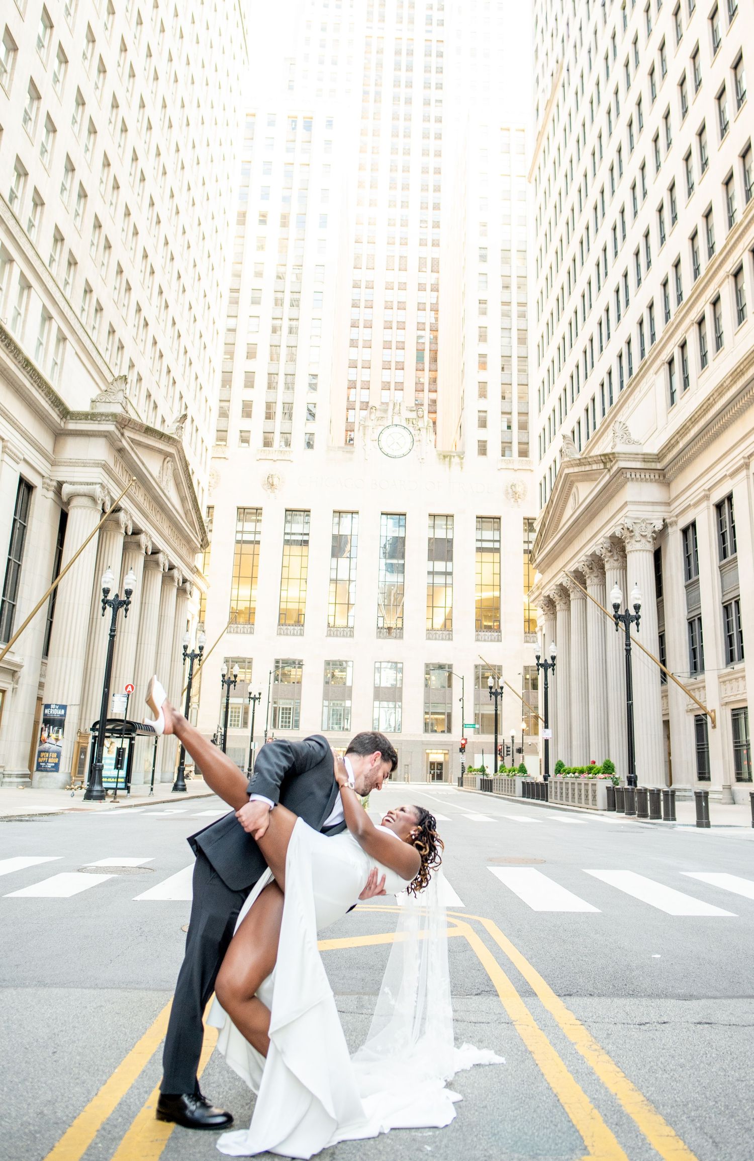 Bride and groom share a romantic dip kiss on a sunlit urban street surrounded by elegant city buildings.