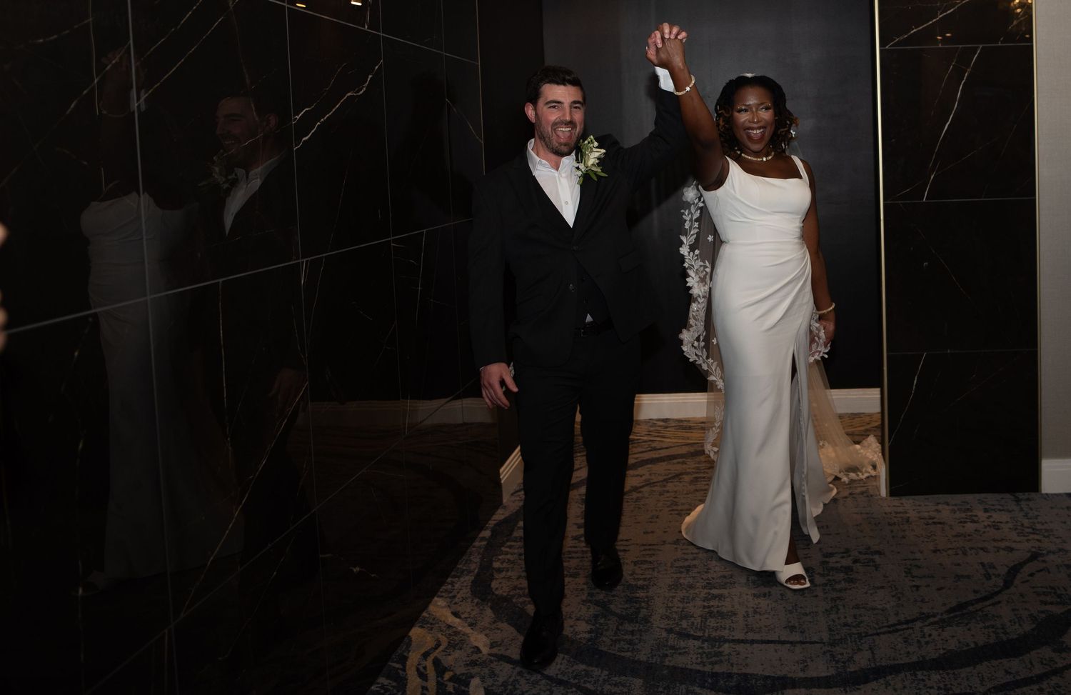 Bride in white gown and groom in black suit wave to guests at elegant wedding reception entrance.