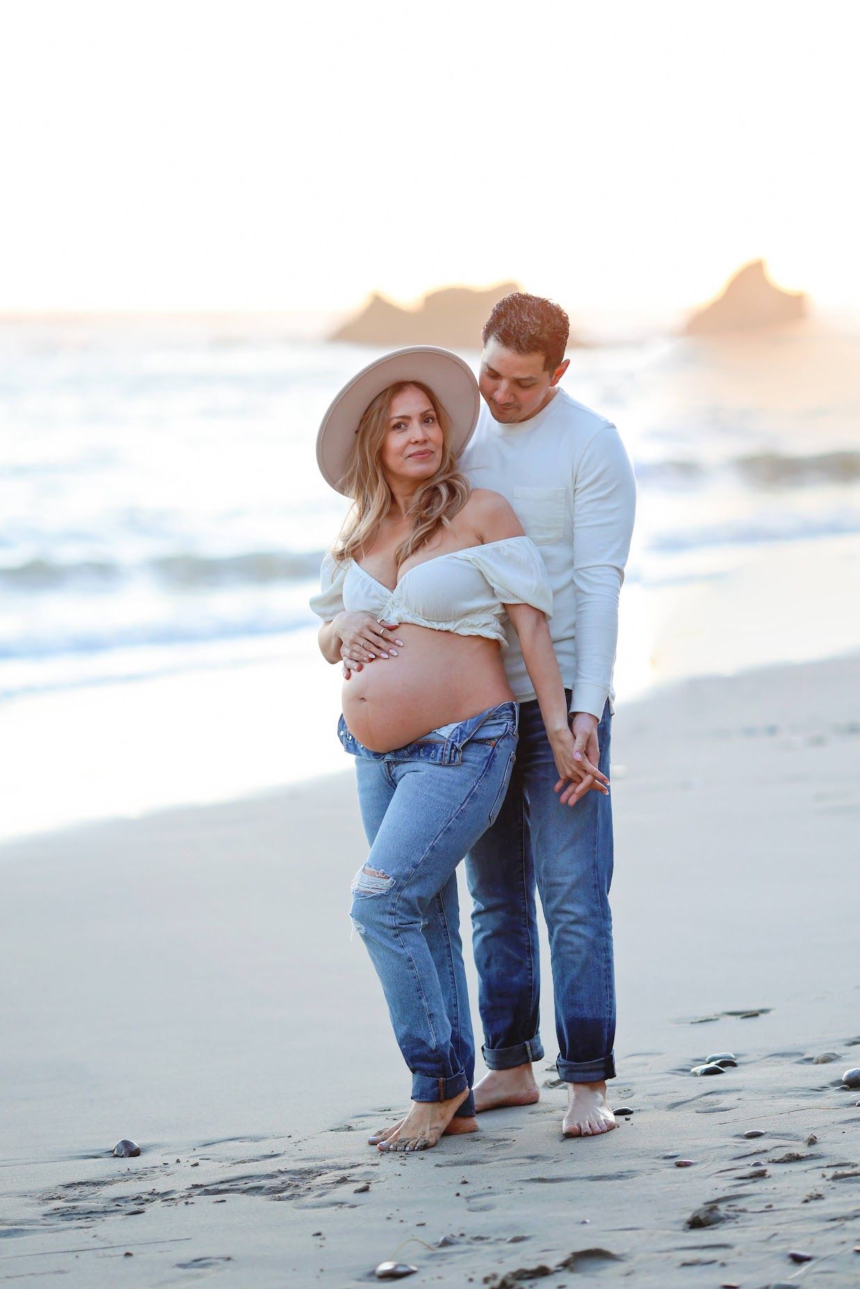 Expecting couple in casual attire embracing on a sandy beach at golden hour sunset maternity shoot.