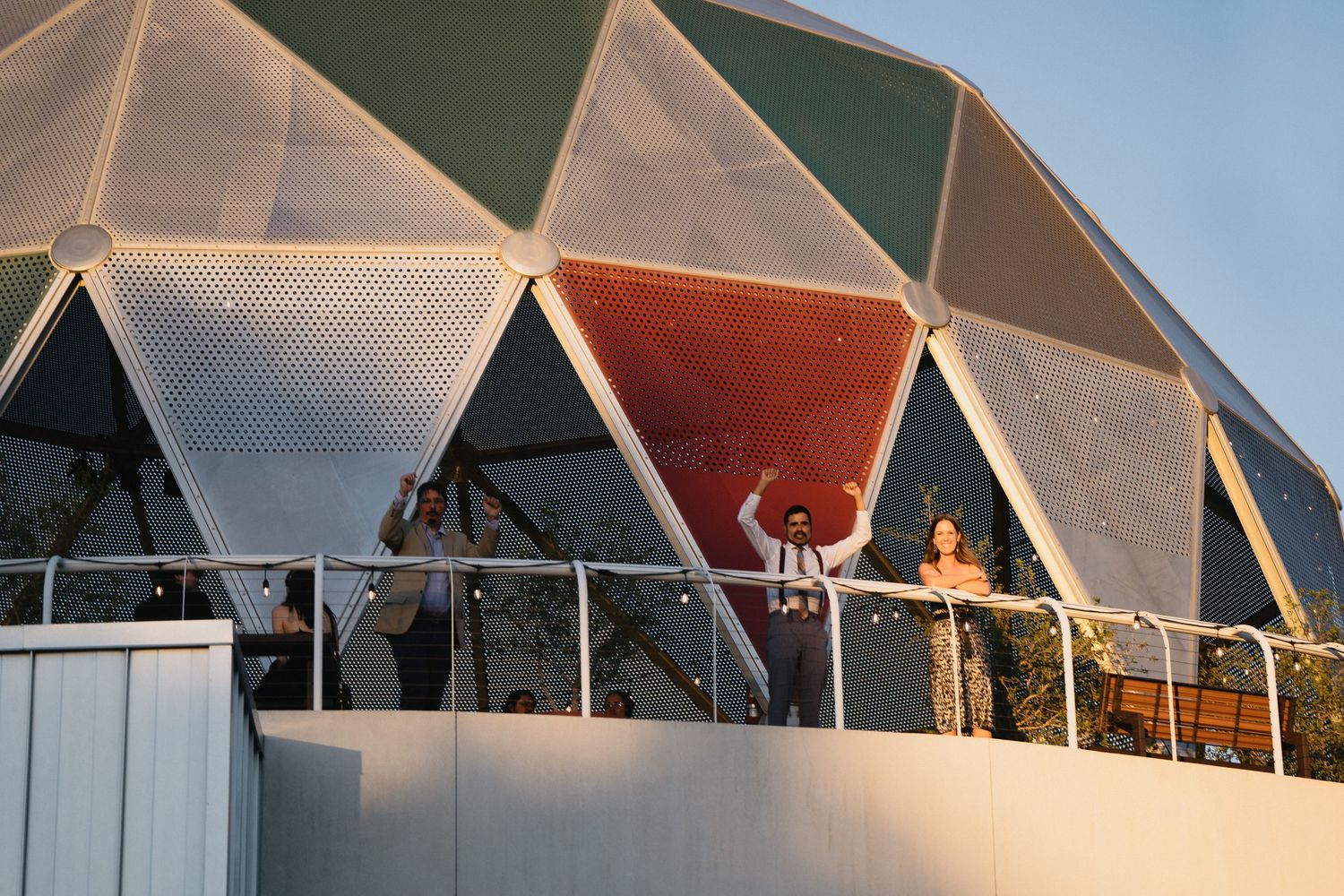 People standing on a balcony in front of a colorful geodesic dome structure outdoors.