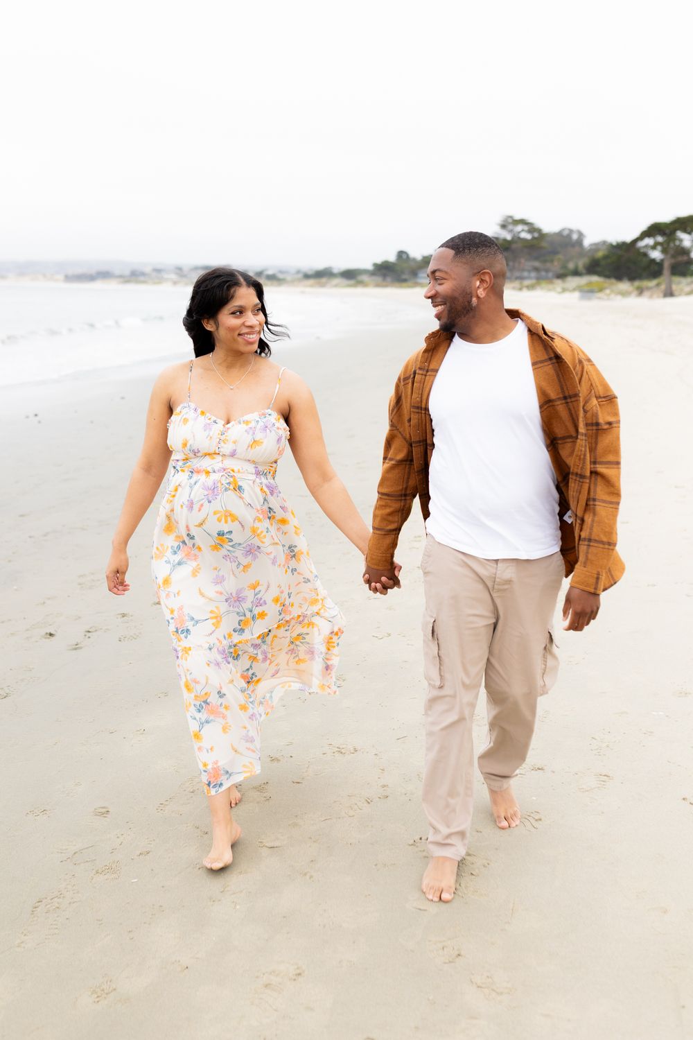 A couple walks hand-in-hand on a beach, smiling at each other, woman in floral dress, man in brown shirt.
