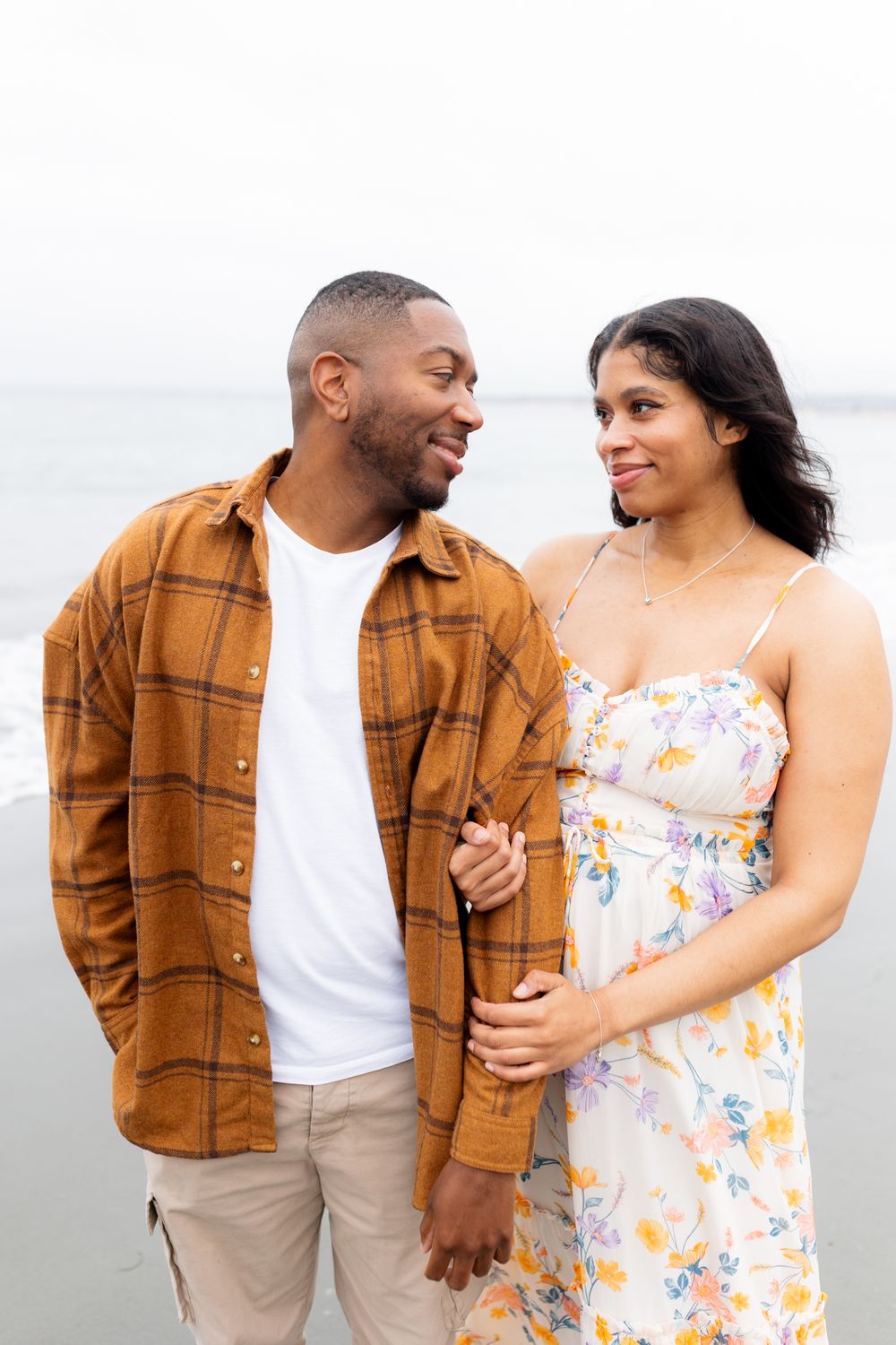 A smiling couple holds hands on a beach, man in plaid shirt, woman in floral dress, sharing a loving glance.