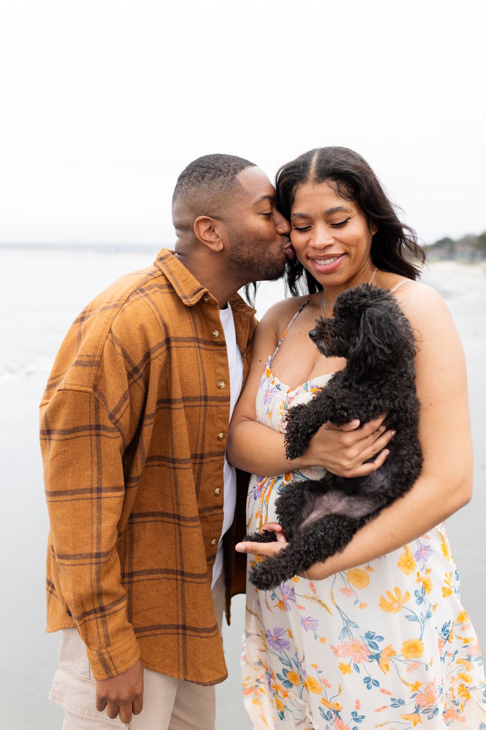 A couple shares a tender moment on the beach, with the man kissing her cheek as she smiles holding a black dog.
