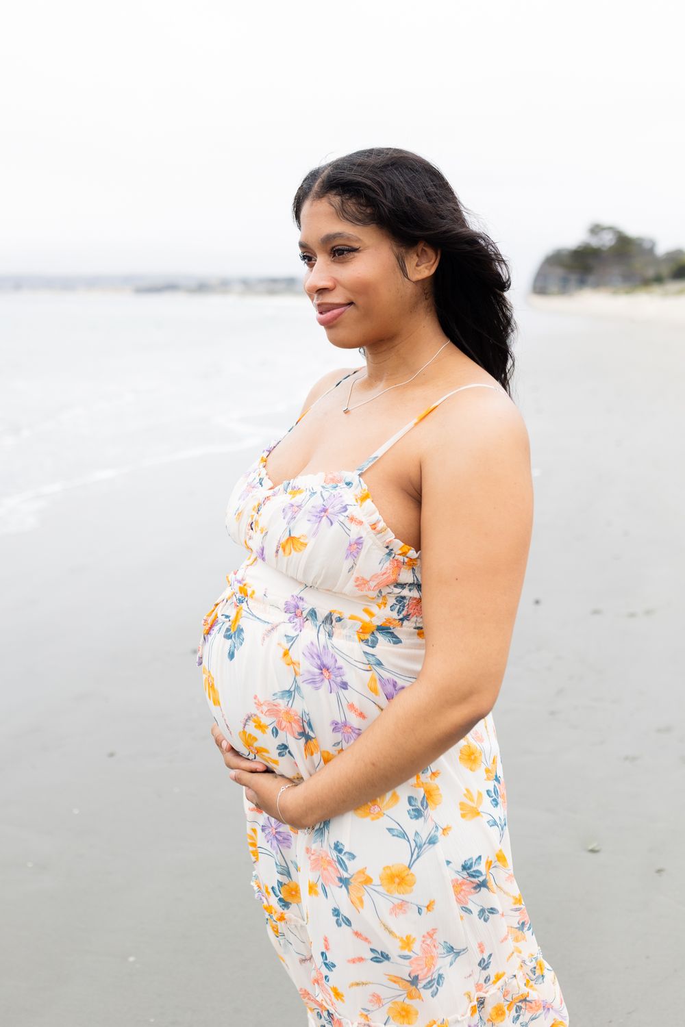 Pregnant woman in floral sundress cradling baby bump on beach, smiling softly with ocean waves behind her.