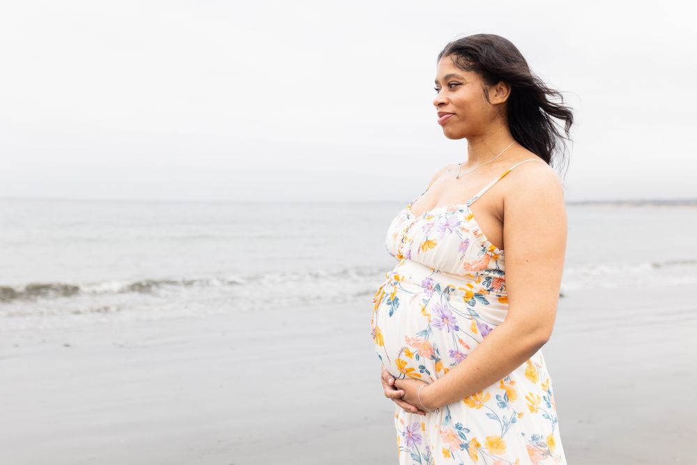 Pregnant woman in floral sundress cradling baby bump on overcast beach, hair blowing in wind.
