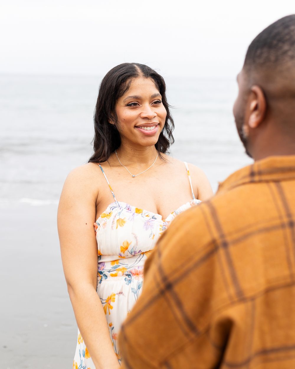 A smiling woman in a floral dress stands on a beach, facing a man in a plaid shirt with the ocean behind her.