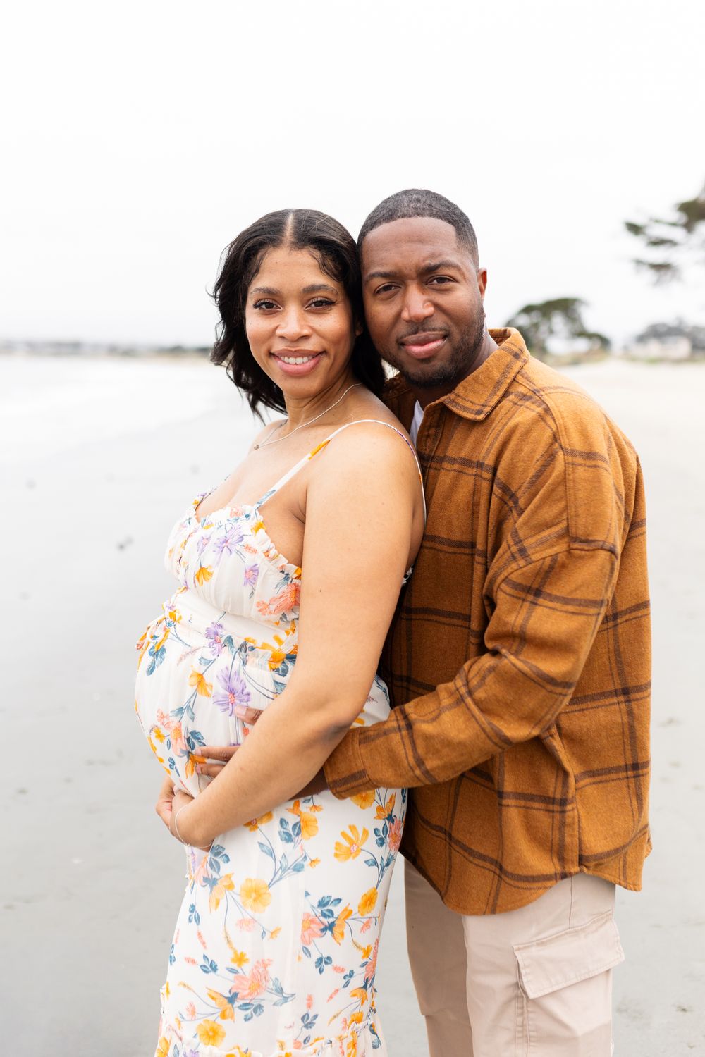 Pregnant couple posing on beach, woman in floral dress cradling baby bump, man in brown plaid shirt embracing her.