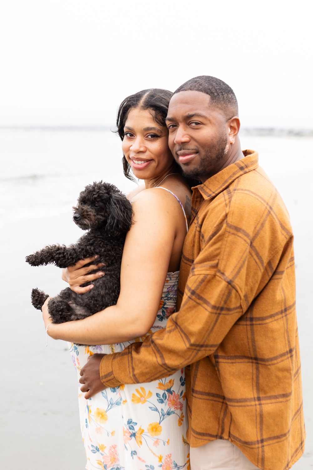 A couple smiles while holding a small black dog on a beach, the woman in a floral dress and man in a plaid shirt.