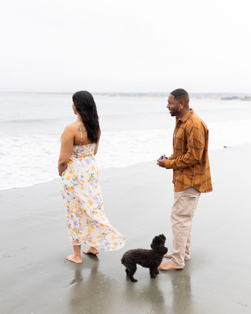 Couple and black poodle standing on a sandy beach near ocean waves, woman in floral dress, man in brown jacket.