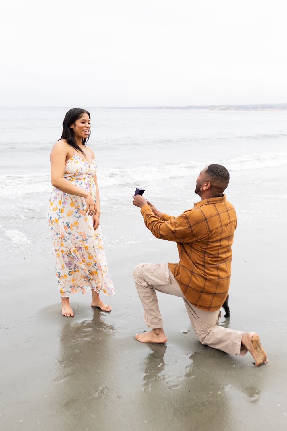 Man proposes on one knee to woman in floral dress on a beach, holding a ring box by the ocean.
