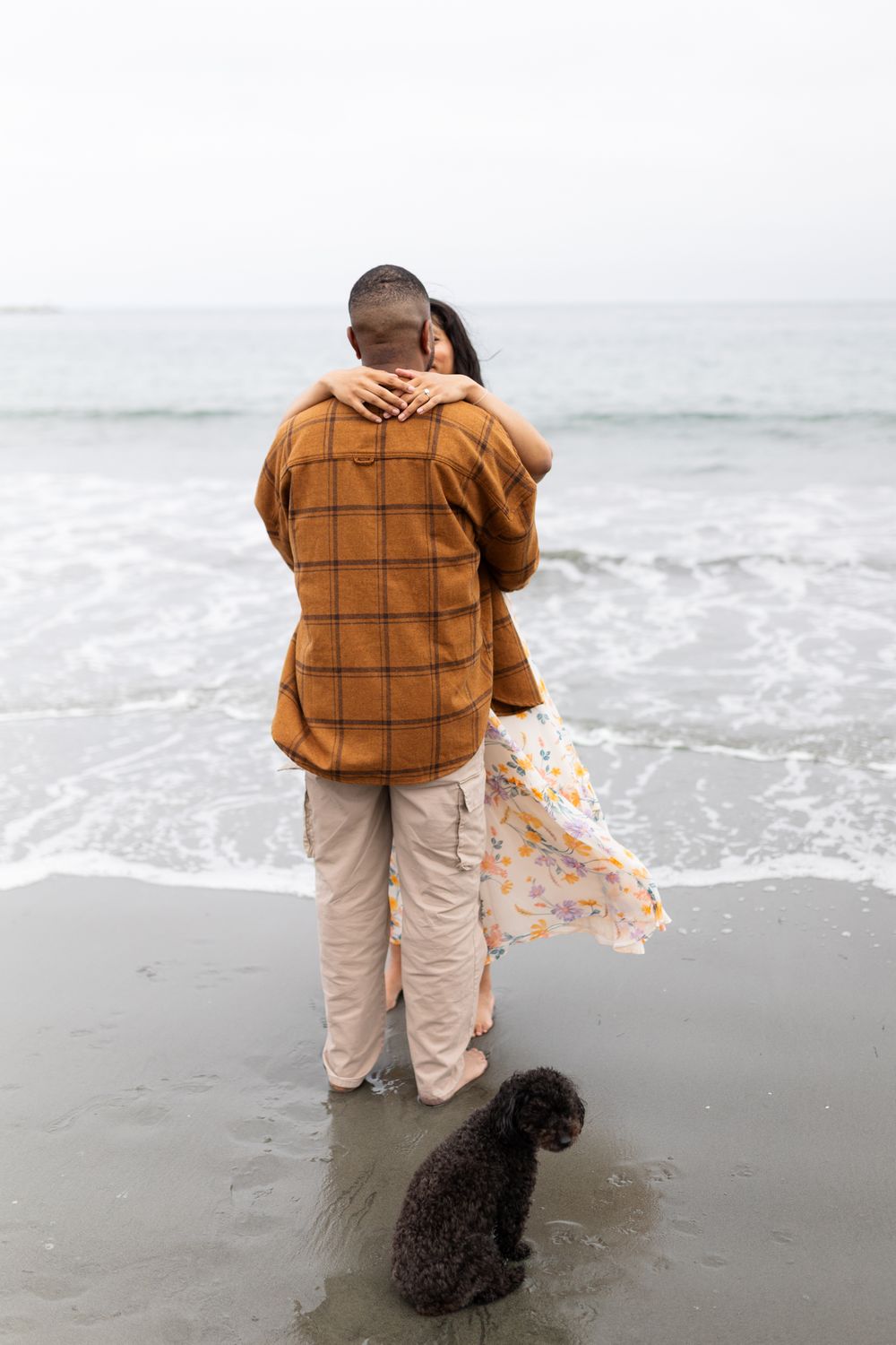 Couple embracing on a beach shoreline with a black dog nearby, wearing casual autumn clothing by the ocean waves.