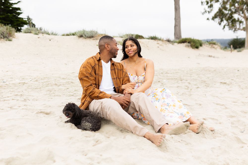 Couple sitting together on sandy beach dunes with their black dog, man in brown jacket, woman in floral dress.