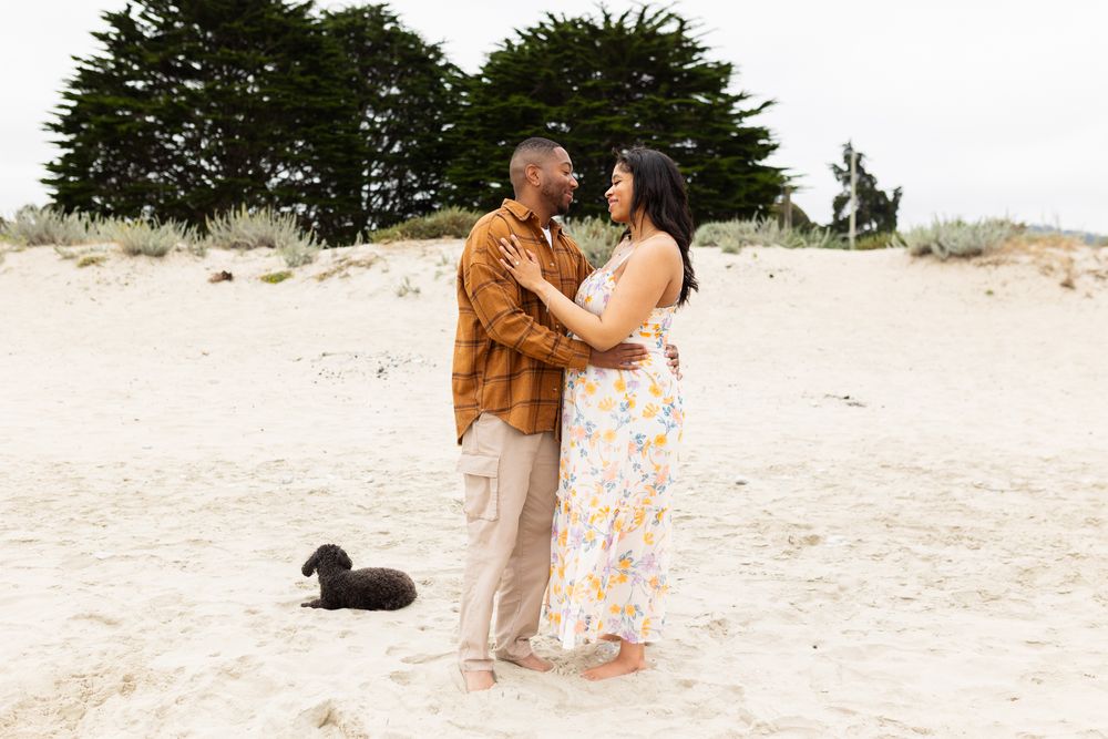 Couple embracing on sandy beach with black dog and cypress trees, maternity photo shoot in natural light.