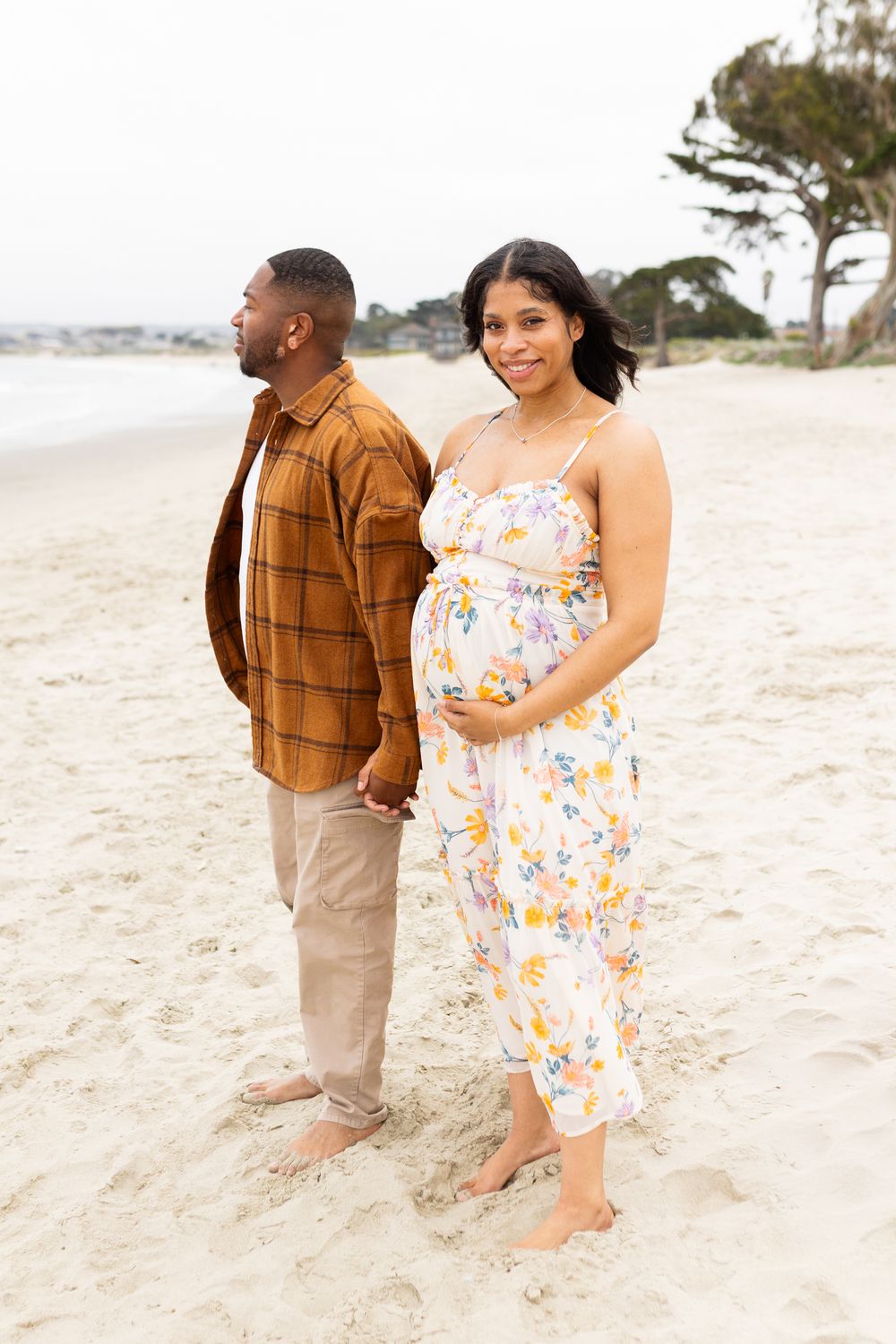Pregnant woman in floral dress holds baby bump on beach beside man in plaid shirt, maternity photo session.