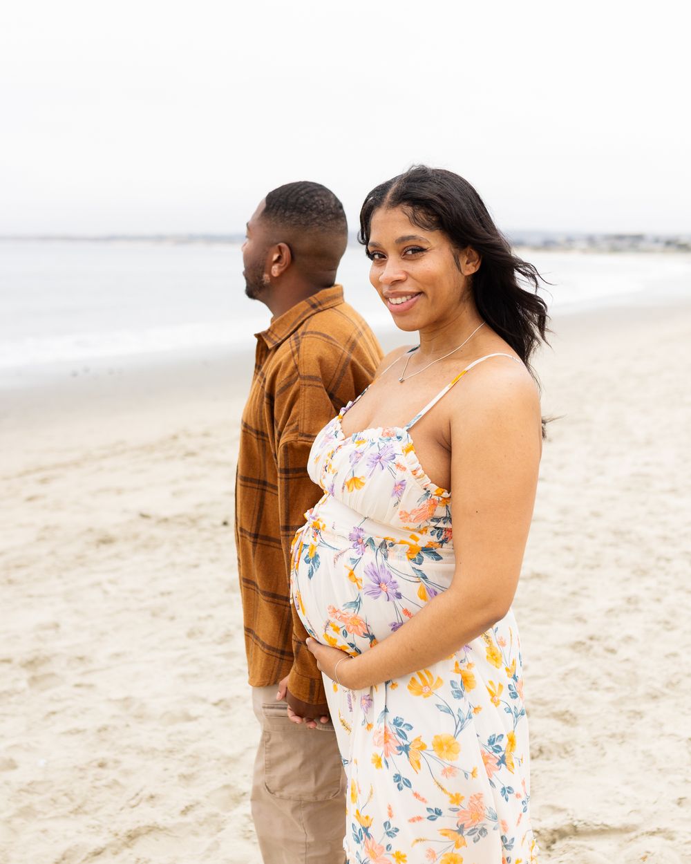 A pregnant woman in a floral dress cradles her baby bump on a sandy beach while her partner stands behind her.