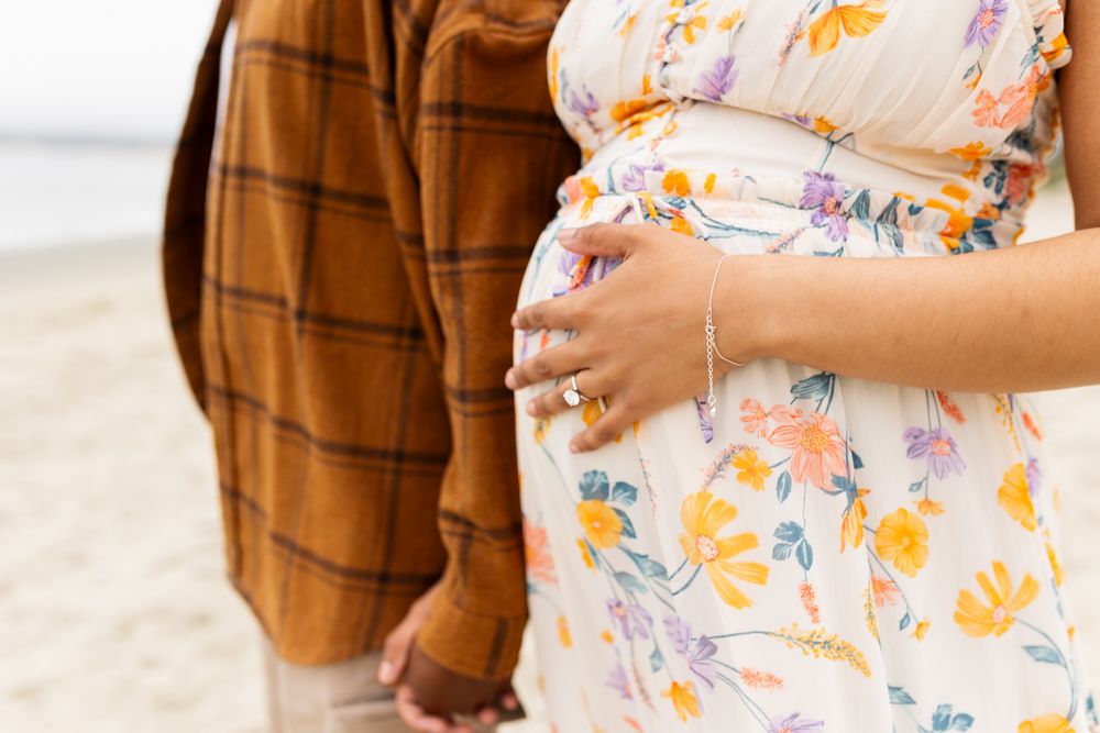 Pregnant woman in floral dress holding baby bump on beach with partner in brown plaid shirt.
