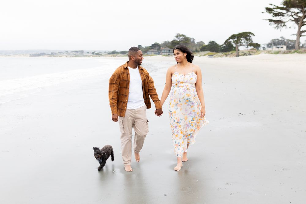 Couple holding hands walking on beach with small black dog, woman in floral dress, man in brown jacket.